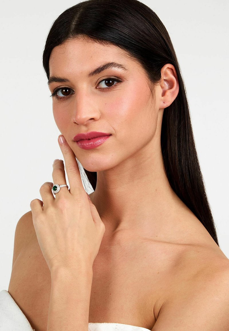 Anello d'argento con una pietra verde ovale, incastonata al centro, circondata da piccole pietre trasparenti. Indossato su una mano appoggiata vicino al viso.
