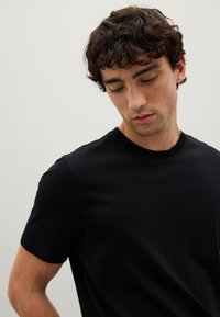 Giovane uomo con capelli ricci e scuri che indossa una semplice maglietta nera a maniche corte, che guarda in basso su uno sfondo neutro.