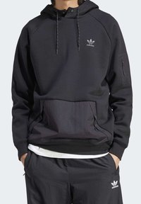 Svart huva sweatshirt med halv dragkedja, som har en framficka och sidodragkedja, tillverkad av ett mjukt material med en adidas-logotyp på bröstet.