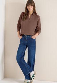 Femme souriante avec les mains dans les poches, portant un chemisier marron à manches longues, un jean bleu à jambes larges et des baskets blanches à rayures vertes.