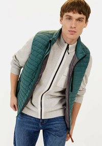 Türkisfarbener gesteppter Weste mit einem Reißverschluss vorne und hohem Kragen, über einem hellgrauen Pullover mit einem weißen und schwarzen Reißverschluss. Sichtbare Jeans aus Denim.