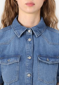Denimskjorte med klassisk krage, frontknapplukking, to brystlommer med klaffer, og en lyseblå farge med en teksturert overflate.