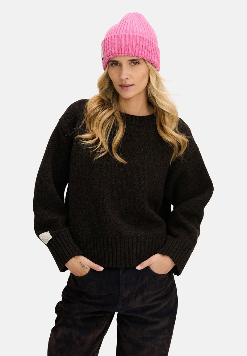 Czarny dziergany sweter z prążkowanym obrzeżem, zestawiony z różową prążkowaną czapką. Model prezentuje luźny fason, ręce w kieszeniach, proste detale projektu.