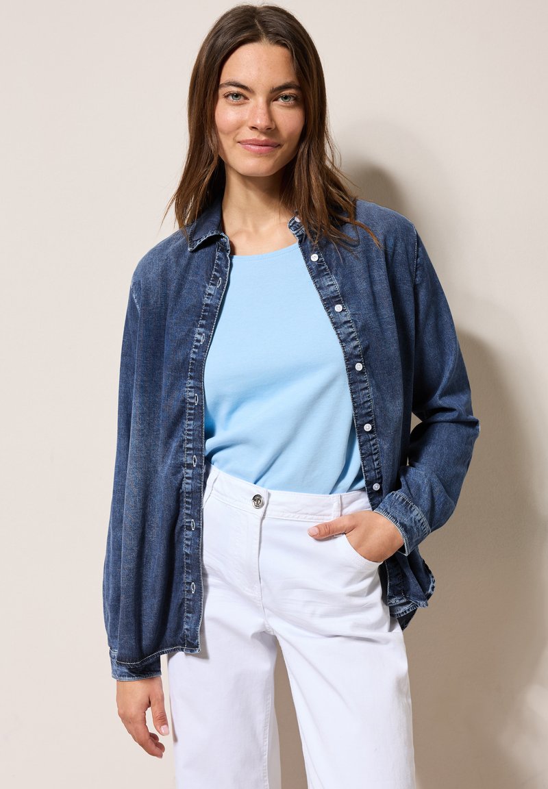 Donkerblauwe denim overhemd met een knoopsluiting en een zachte textuur; gecombineerd met een lichtblauw t-shirt en witte broek.