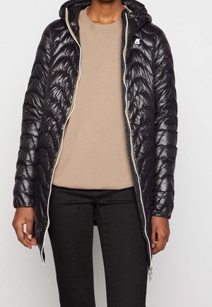 Personne portant une longue veste matelassée noire à capuche par-dessus un pull beige et un pantalon noir, debout les bras détendus le long du corps.