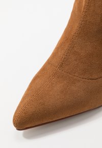 Gros plan sur le bout pointu d'une botte cheville en daim marron avec des coutures visibles sur un fond blanc.