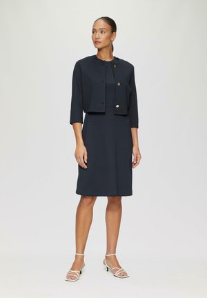 Vestido azul marino con una silueta ajustada y dobladillo a la altura de la rodilla, acompañado de una chaqueta corta con acentos en botones dorados. Sandalias blancas con tiras completan el look.
