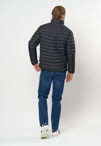 Man med en svart vadderad jacka, blå jeans och vita sneakers, stående och gående bortåt mot en enkel vit bakgrund.