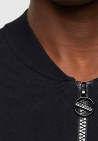 Giacca bomber nera in maglia con colletto a coste, dotata di zip visibile e cursore rotondo con il marchio "DIESEL".