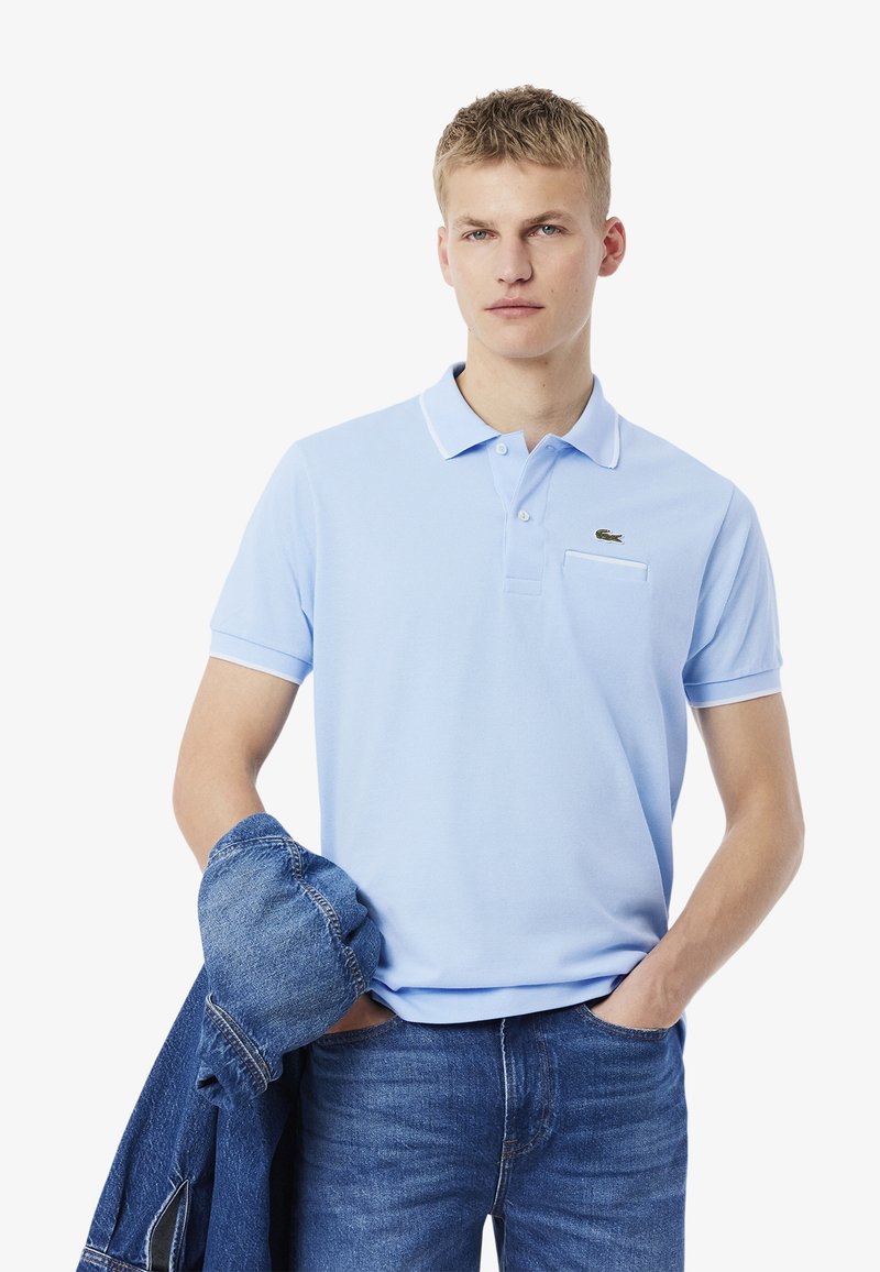 Ljusblå polo med krage, ficka och grön logotyp. Bärs med blå denimjeans och en denimjacka över armen.