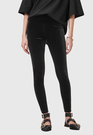 Leggings in velluto nero con un design elegante, vita alta e una texture liscia, abbinati a scarpe basse con cinturini neri e dorati.