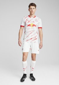 Balta futbolo apranga su raudonomis detalėmis, turinti įstrižainėmis juostelėmis raštą ir išsiskiriantį logotipą. Pakuotėje yra šortai, iki kelių siekiančios kojinės ir futbolo batai.
