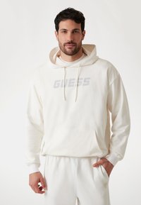 Weißer Hoodie mit Netzstruktur, Fronttasche und „GUESS“-Logo in Hellgrau. Mit Kordelzugkapuze und gerippten Bündchen, kombiniert mit weißen Hosen.