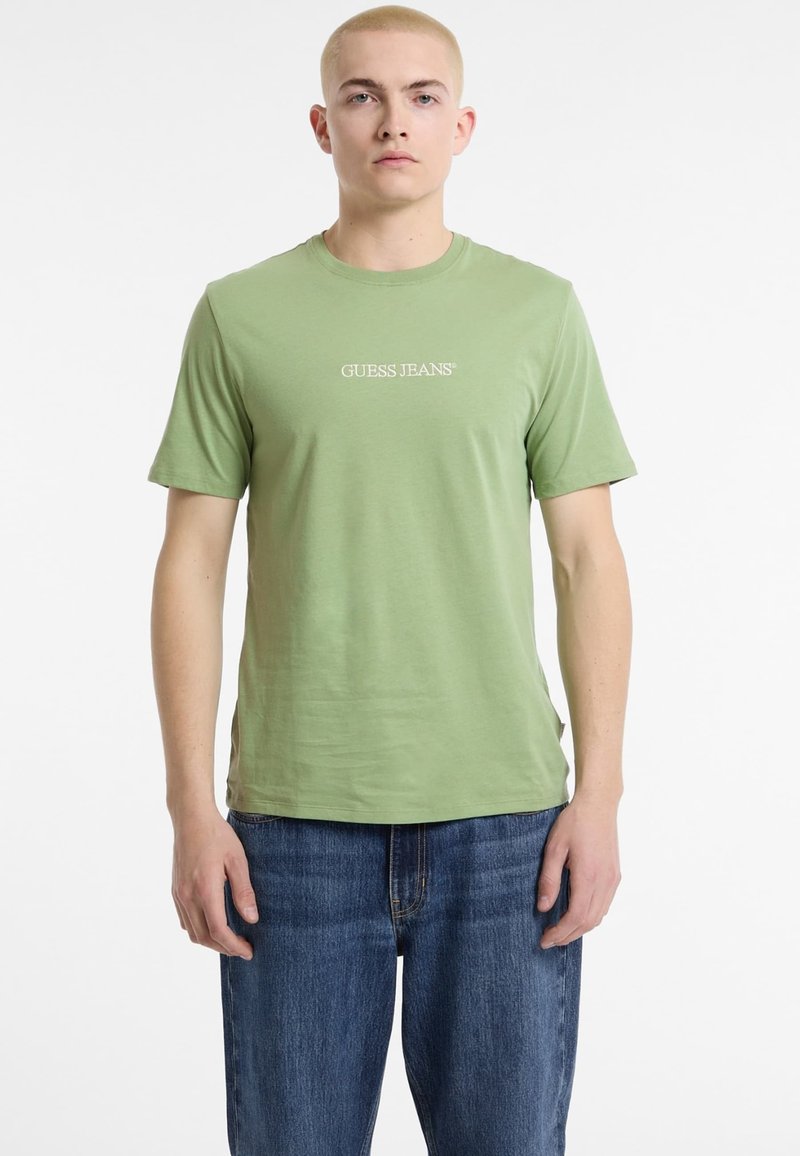 Roheline puuvillane T-särk ümarate kraega, lühikeste varrukatega ning valge "GUESS JEANS" logo rinnal, millele on sobitatud sinised teksapüksid.