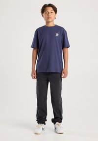 Chico adolescente de pie, vistiendo una camiseta gráfica azul marino, jeans negros y zapatillas Nike blancas, contra un fondo liso claro.