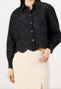 Black, short-sleeved button-up shirt, eyelet embroidery with scalloped hem and cut-out patterns; paired with a light-colored ribbed skirt.