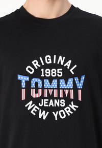 Sort bomulds T-shirt med hvid tekst "ORIGINAL 1985 TOMMY" med røde striber og blå sektioner med stjerner. "JEANS NEW YORK" under.