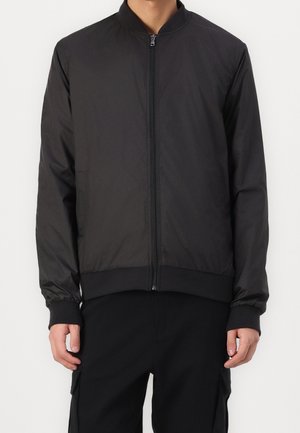 Homme portant une veste bomber noire unie à fermeture éclair, avec poignets et taille côtelés, associée à un pantalon noir sur fond blanc.