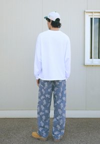 Osoba stojící zády k fotoaparátu, oblečená v bílé košili s dlouhým rukávem, vzorovaných volných džínách, hnědých botách a bílé čepici, vedle šedé zdi s oknem.