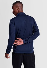 Marinblå, långärmad atletisk jacka med hög krage, slät textur och åtsittande ärmslut, som bärs över svarta byxor med avslappnad passform.