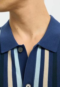 Polo majica z naramnicami z rastrami temno modre, svetlo modre in bež barve. Klasičen ovratnik z dvema gumboma in teksturirano blago.