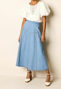 Jupe midi en denim bleu avec une silhouette évasée, des détails de couture verticale et une taille haute. Associée à une blouse blanche et des talons compensés.