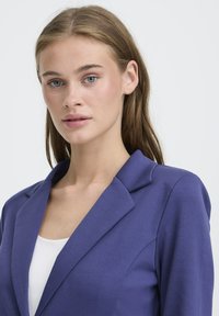Mlada ženska z svetlo rjavimi lasmi in modrimi očmi, oblečena v modro blazer jakno in belo majico, ki se spoprijateljuje s pogledom proti naprej na enobarvnem svetlem ozadju.
