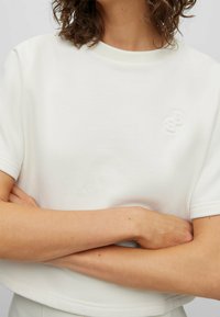 Kobieta w prostym, białym, krótkim topie crop z małym wytłoczonym logo na górnej lewej części klatki piersiowej, z rękami skrzyżowanymi na piersiach.