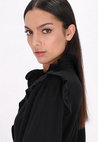 Blusa nera con colletto alto e particolari arricciati, dettaglio dei bottoni e tessuto testurizzato. Maniche lunghe con vestibilità comoda.