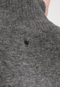 Pull en tricot gris avec un col haut, présentant un petit accent métallique. La texture est douce et moelleuse, avec des fibres visibles et un motif côtelé.
