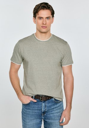 Młody mężczyzna o jasnej karnacji, ubrany w szarą koszulkę z krótkim rękawem i niebieskie dżinsy, stojący z jedną ręką w kieszeni na jednolitym tle.