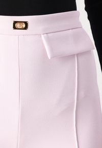Saia rosa clara feita de tecido texturizado, apresentando um detalhe em laço na frente e um fecho de metal dourado com logotipo em relevo. Acabamento suave.