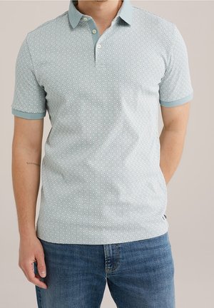 Homme portant un polo manches courtes bleu clair à motifs avec boutons et un jean bleu, debout devant un fond neutre.