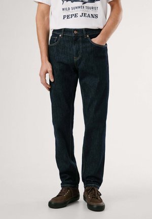 Mand iført mørkeblå jeans med lige ben, brune sneakers og en hvid T-shirt med grafik og teksten "Wild Summer Tourist Pepe Jeans."