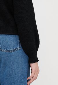Pulôver de malha preto com uma textura aconchegante e mangas longas ligeiramente volumosas, combinado com jeans azul de cintura alta.