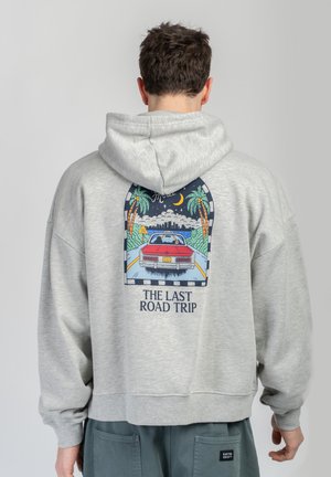 Osoba w szarej bluzie z kapturem z kolorową grafiką na plecach przedstawiającą czerwony samochód na drodze obsadzonej palmami oraz napisem "The Last Road Trip".