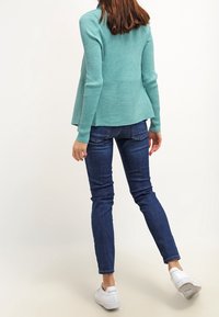Suéter de punto acanalado en turquesa con dobladillo de peplum, combinado con jeans ajustados azul oscuro y zapatillas blancas, visto desde atrás.
