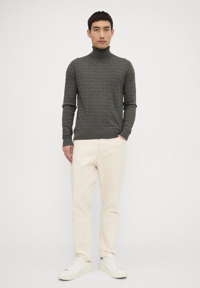 Maglione grigio a collo alto con un motivo a logo ripetuto, abbinato a pantaloni chinos color crema e sneakers bianche, in piedi contro uno sfondo neutro.