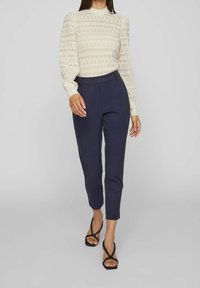 Valkoinen kuvioitu pitkähihainen paita korkealla kaula-aukolla, yhdistettynä tummansinihin räätälöityihin housuihin ja mustiin remmikenkiin. Sileä pinta, istuva malli.