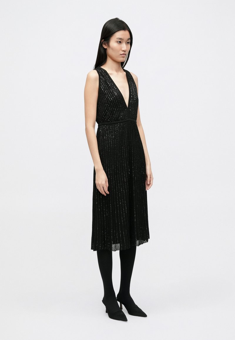 Black dress with a deep V-neckline, pleated texture, and shimmering stripes. Paired with black tights and pointed-toe heels.