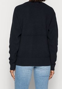 Černý pletený svetr s ribovanou strukturou a lemem, s kleslými rameny. Nosený s světle modrými džíny.