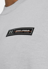 Grå t-shirt med en struktureret stof, der har et sort mærke med hvid og orange tekst. Rund halshulm, korte ærmer.