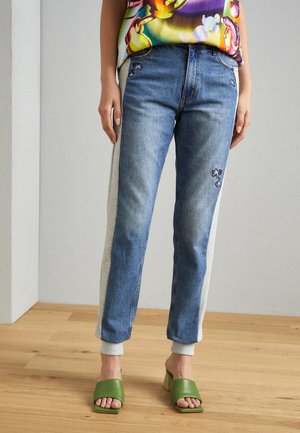 Slim-fit jeans met grijze zijpanelen en bloemrijke borduursels, gecombineerd met groene open-toe blokhakken op een houten vloer.