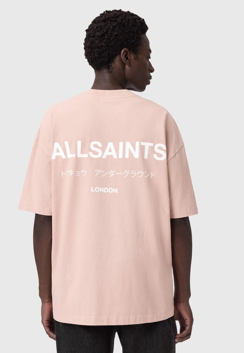 Rövid hullámos hajú férfi, laza, rózsaszín pulóvert visel, amelyen fehér „ALLSAINTS” felirat és japán szöveg található a hátoldalon, háttal a kamerának.