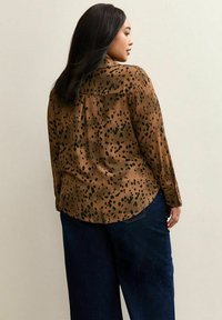 Femme aux longs cheveux bruns portant une chemise marron à boutons avec des taches noires et un jean large bleu foncé, debout contre un mur uni.