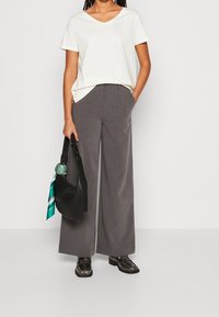 Camiseta blanca de cuello en V con adornos verticales, combinada con pantalones de pierna ancha grises. Bolso negro con bufanda de estampado verde, zapatos negros.