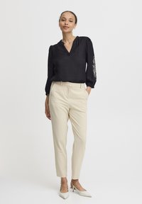 Blusa nera con maniche a sbuffo e scollatura a V, decorata con ricami floreali bianchi su una manica. Abbinata a pantaloni tapered beige e tacchi bianchi.