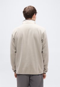 Μπεζ πουλόβερ φλις με ψηλό γιακά, μακριά μανίκια, ελαστικές μανσέτες και λεία υφή. Χωρίς ορατά λογότυπα ή σχέδια. Πλάγια όψη από πίσω.