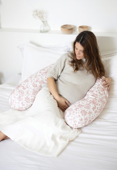 Μαξιλάρι εγκυμοσύνης με ροζ λουλουδάτο σχέδιο σε λευκό ύφασμα, μακρύ και στρογγυλεμένο σχήμα, μαλακή υφή, χρησιμοποιείται από άτομο σε άνετο κρεβάτι.