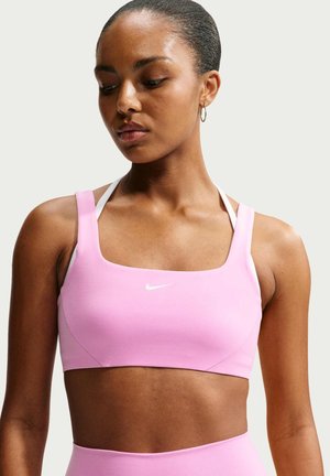 Žena nosi svetloružovú športovú podprsenku s bielymi ramienkami a malým logom Nike, pozerá sa dole na jednoliate pozadie.